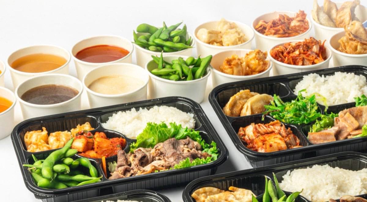 KPOT 2 GO bento boxes packed with beef, rice, kimchi and edamame beside cups of soup and dipping sauces