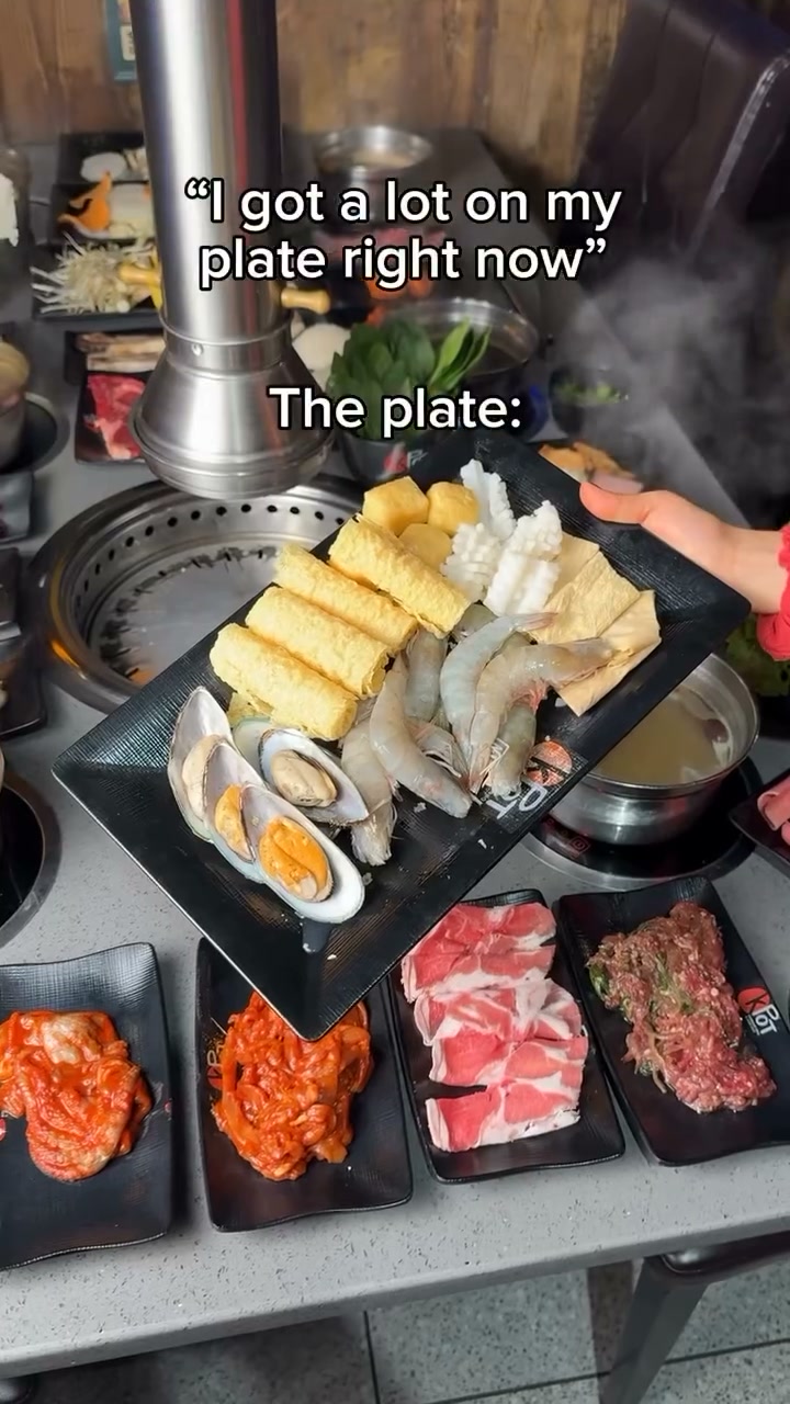 A platter of raw shrimp, mussels, squid, fish cake and tofu puffs held over a KPOT grill, with plates of kimchi and marinated beef below